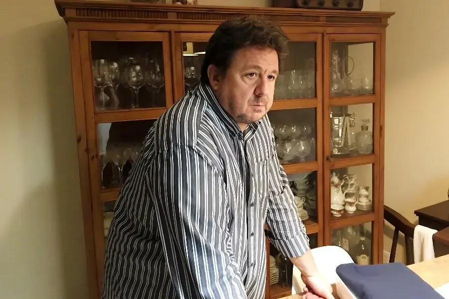 Un hombre con una camisa a rayas está de pie, apoyado en una mesa de madera frente a una vitrina llena de vajilla y cristalería; su expresión seria deja entrever la gravedad del caso Gürtel.