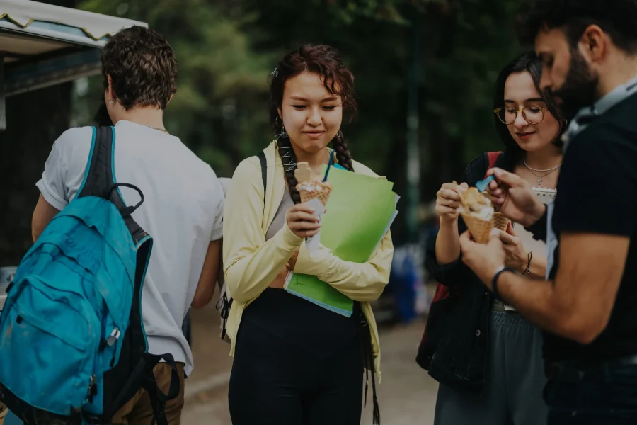 Cuatro jóvenes están al aire libre; tres de ellos disfrutan de unos helados en cucurucho, mientras que otro, con una mochila y una carpeta verde —quizá un «Borrador automático»—, se une a la conversación. Los frondosos árboles crean un agradable telón de fondo mientras disfrutan juntos de sus helados.