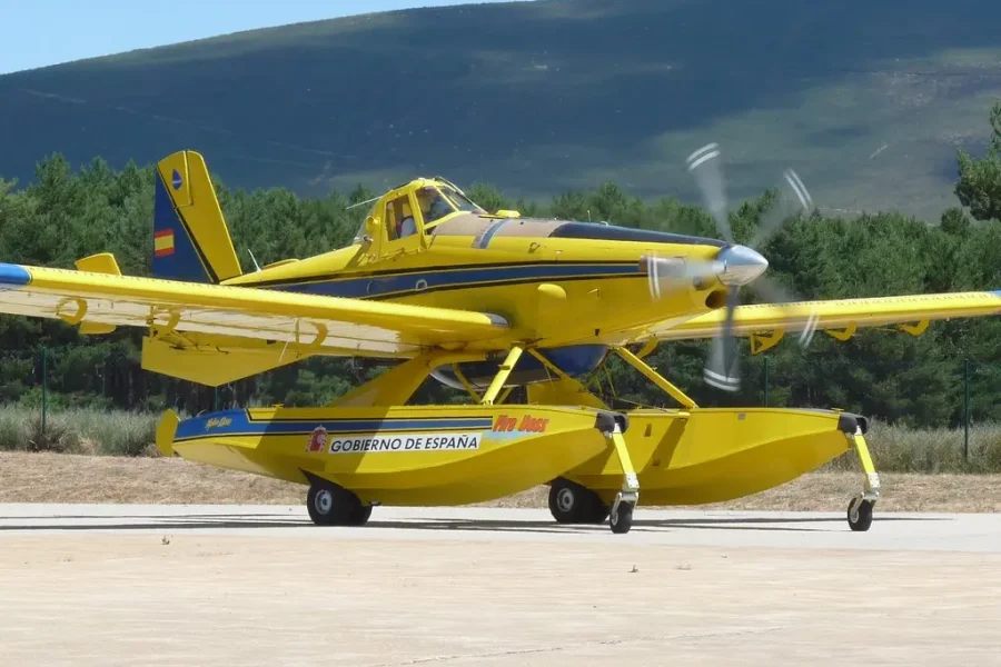 Un avión anfibio amarillo con pontones y la inscripción «Gobierno de España» en el costado, identificado como «borrador automático», está aparcado sobre una superficie de hormigón, con un bosque y una colina al fondo.