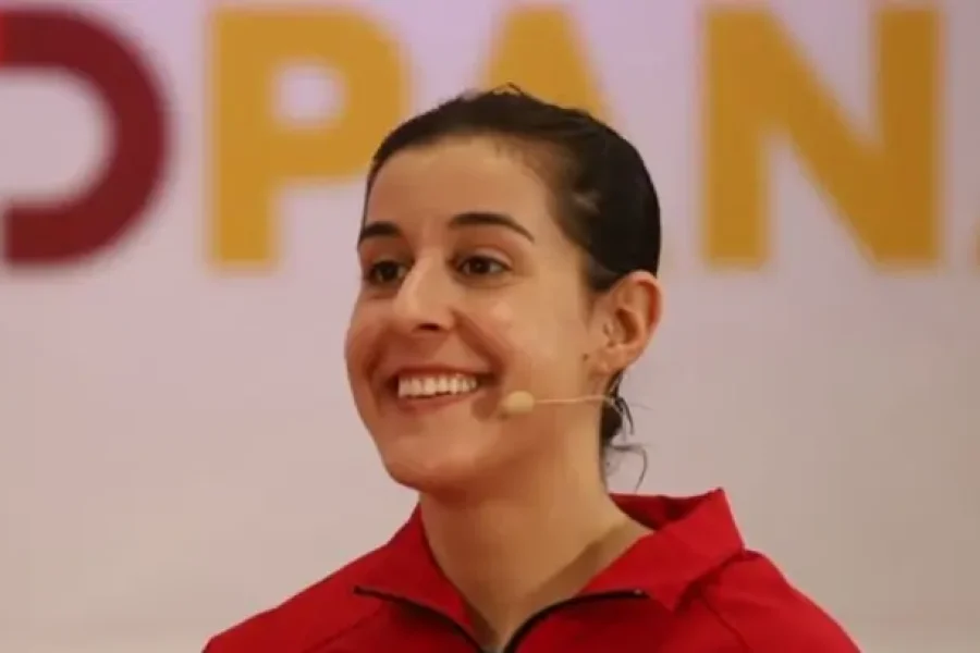 Una persona que lleva una chaqueta roja y unos auriculares con micrófono está sonriendo, con un aspecto muy parecido al de Carolina Marín durante la ceremonia de entrega del Premio al Mérito Deportivo. En el fondo, difuminado, se ven unas letras grandes, parcialmente visibles, de color amarillo y rojo.