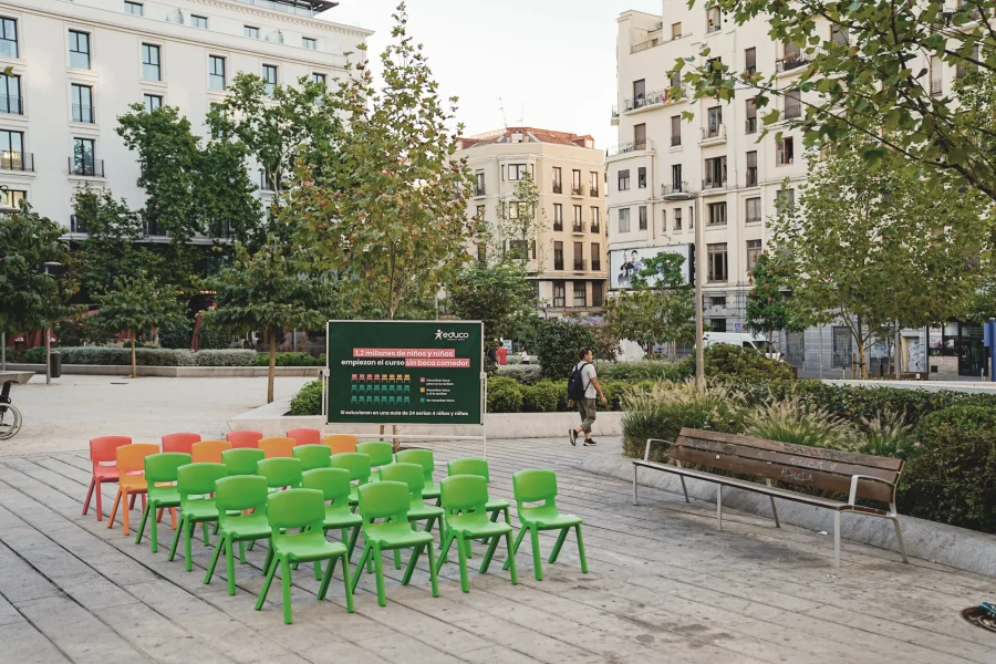 Varias filas de sillas de plástico verdes y naranjas están orientadas hacia un gran cartel situado en una plaza urbana, rodeada de árboles y con varios edificios de varias plantas al fondo. Una persona pasa por allí, quizá reflexionando sobre programas como las becas de comedor escolar, mientras que cerca hay un banco de madera.