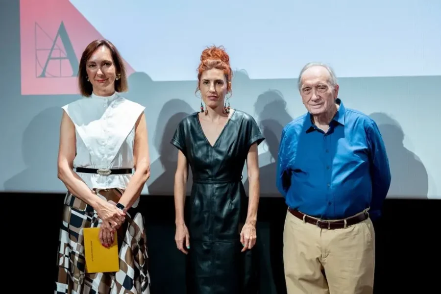 Tres adultos, que participan en el evento «Óscar España» de preselección, posan delante de una pantalla de proyección. La mujer de la izquierda lleva un top blanco y una falda estampada; la mujer del centro, un vestido negro; y el hombre de la derecha, una camisa azul y unos pantalones beige.