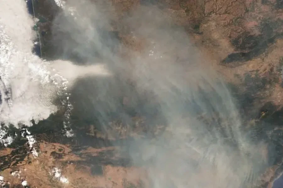 Una imagen de satélite muestra el humo de los incendios forestales en España extendiéndose por un paisaje seco, con nubes a la izquierda y elementos del terreno visibles a través de la neblina. En la esquina superior derecha se aprecian la costa y una gran masa de agua.
