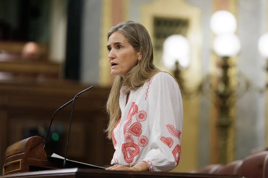 Una mujer de pelo largo, vestida con una blusa blanca con bordados rojos, habla desde un atril en una sala ornamentada con iluminación decorativa y paneles de madera al fondo, refiriéndose a la reciente ola de calor que ha marcado el verano más cálido en España.