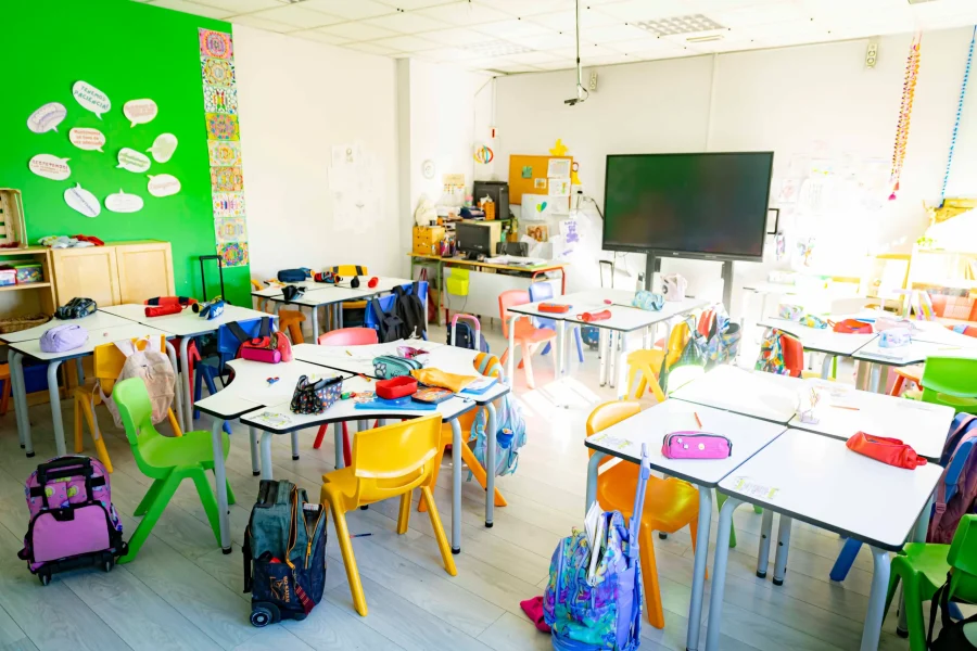 Un aula bien iluminada en colegios públicos, con pupitres blancos, sillas de colores, mochilas y estuches de lápices. Una pared verde con carteles, una gran pantalla en la parte delantera y material diverso decoran este espacio de aprendizaje bien equipado.