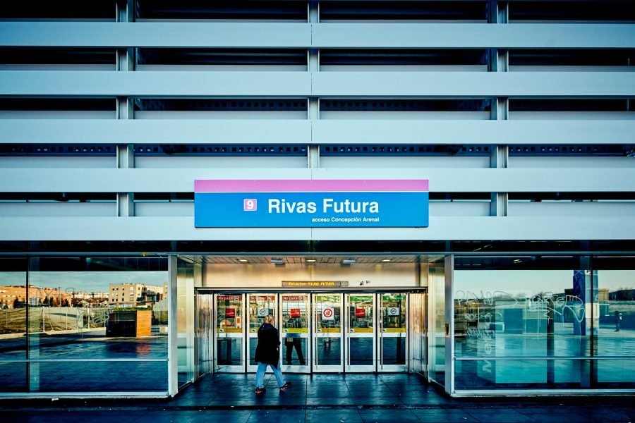 Entrada de la estación de Rivas Futura: bajo el cartel azul con la banda rosa y el logotipo de Metro de Madrid, la puerta acristalada de la estación y una persona caminando, en una imagen nocturna.
