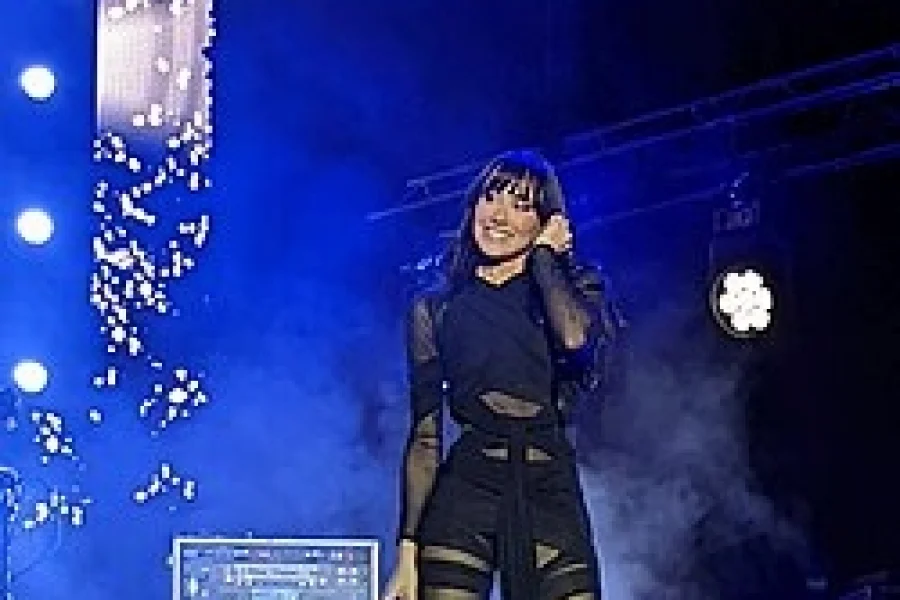 Aitana, vestida de negro, actúa en el escenario con una sonrisa, sosteniendo un micrófono mientras las luces azules del escenario y el humo crean una atmósfera eléctrica de fondo.
