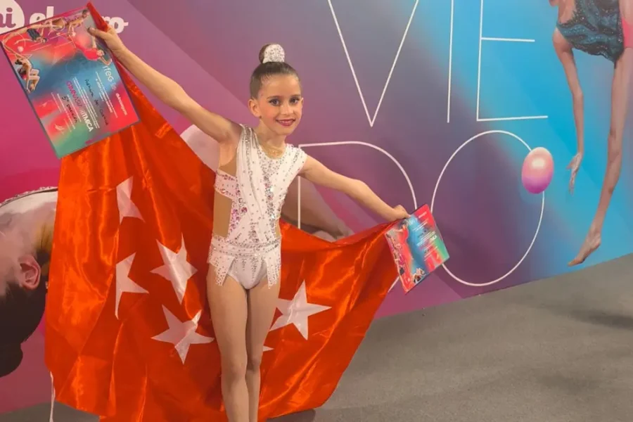 Adhara Rivas, una joven con un maillot blanco, sostiene una gran bandera roja con estrellas blancas y dos certificados. Se alza orgullosa ante un colorido telón de fondo con grandes letras, imágenes de gimnastas y símbolos de gimnasia rítmica.