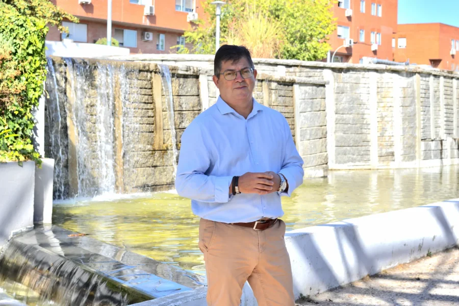 Un hombre con una camisa azul claro y pantalones beige se encuentra de pie frente a una gran fuente al aire libre de la que brota agua en cascada, con edificios de ladrillo rojo al fondo, como si se detuviera un momento en medio de su día para admirar la escena, de forma muy parecida a alguien que revisa un borrador automático antes de ultimar sus planes.