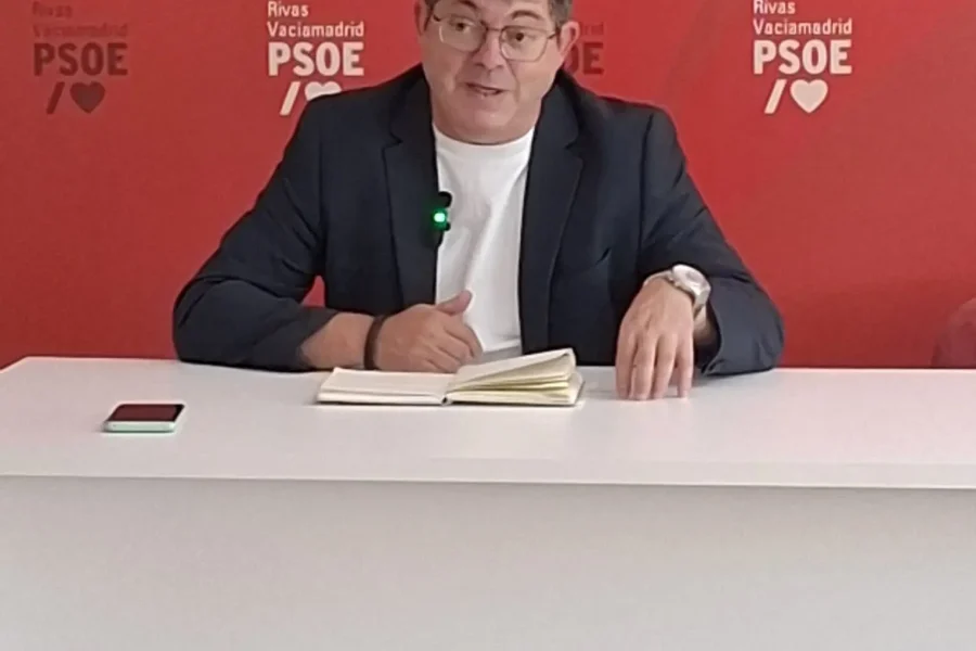 Un hombre con gafas y chaqueta está sentado ante un escritorio blanco, con un portátil y un teléfono, hablando del «borrador automático» frente a un fondo rojo con los logotipos del PSOE de Rivas Vaciamadrid.