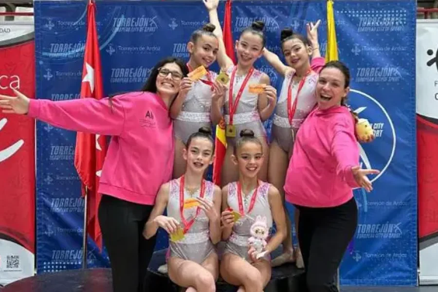 Seis jóvenes gimnastas alevines con leotardos y dos mujeres con sudaderas rosas posan juntas, sonrientes y sosteniendo medallas, celebrando como campeonas de Madrid en una competición de gimnasia en Rivas.