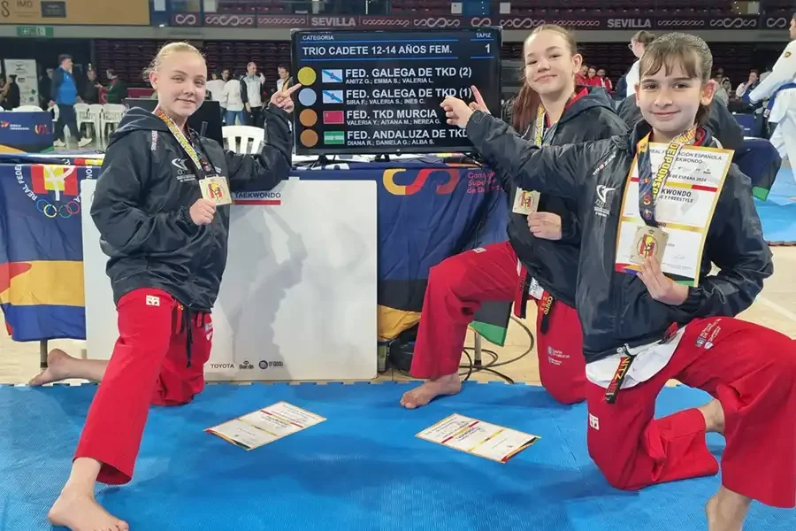 Tres jóvenes artistas marciales vestidas con pantalones rojos y chaquetas negras se arrodillan sobre una colchoneta azul, sosteniendo medallas y certificados, delante de un marcador de competición en el que figuran los equipos y los resultados. Al fondo, otros competidores y jueces.