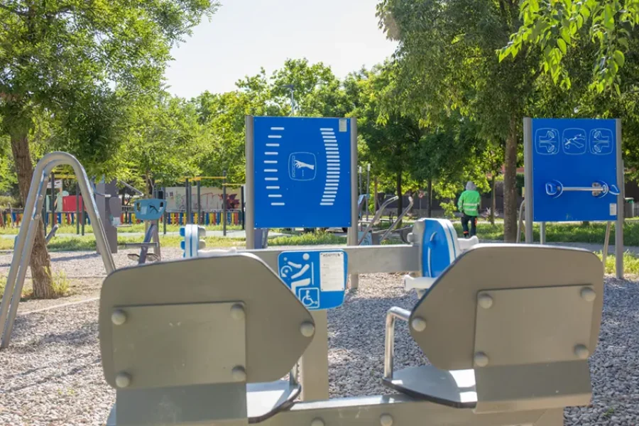 En un parque con árboles frondosos hay un conjunto de aparatos de gimnasia al aire libre instalados sobre una superficie de grava. En unos paneles informativos azules se muestran diagramas de ejercicios de «Borrador automático». Al fondo se ve a una persona con una camiseta verde.