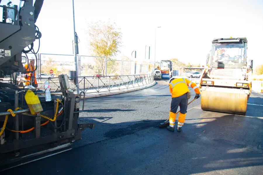 Un trabajador de la construcción de carreteras con uniforme reflectante utiliza un borrador automático para alisar asfalto fresco mientras una apisonadora y otra maquinaria operan cerca en un día soleado.