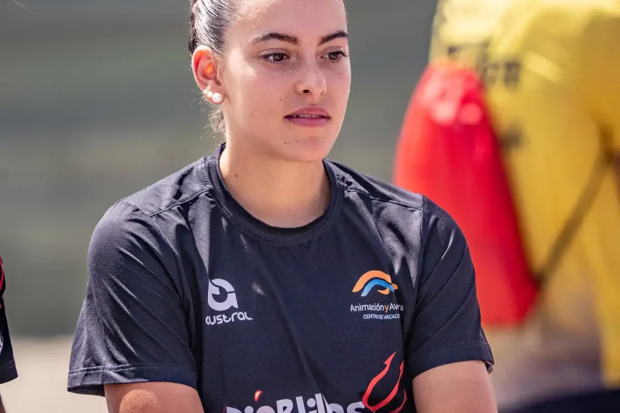 Una mujer joven con el pelo trenzado está de pie con los brazos cruzados, llevando una camiseta deportiva oscura con los logotipos de Diablillos de Rivas y Triatlón, junto con una medalla alrededor del cuello. En el fondo borroso se ven otras personas.