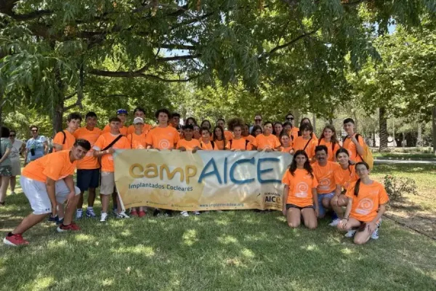 Un grupo de jóvenes, vestidos con camisetas naranjas a juego, están de pie al aire libre sobre el césped en Granada, sosteniendo una pancarta amarilla y azul en la que se lee «CAMP AICE - Implante Coclear». Se encuentran bajo unos árboles frondosos en un día soleado.