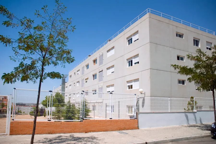 Un moderno bloque de pisos rectangular, con paredes exteriores blancas y hileras de ventanas, situado cerca de un parque público, está rodeado por una valla metálica blanca y un pequeño patio con árboles bajo un cielo azul claro.