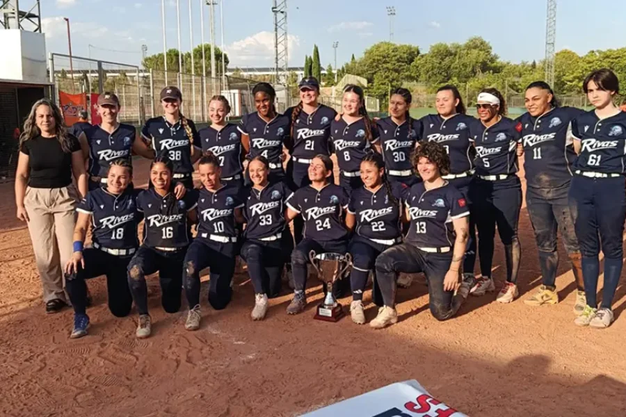 Un equipo femenino de sóftbol, con uniformes azul marino a juego y la inscripción «Riders» en el pecho, posa al aire libre en un campo de tierra. Están reunidas alrededor de un trofeo, algunas arrodilladas y otras de pie, con una mujer de pie en el extremo izquierdo. En representación del CBS Rivas, su unidad y orgullo se hacen evidentes mientras celebran juntas su logro.