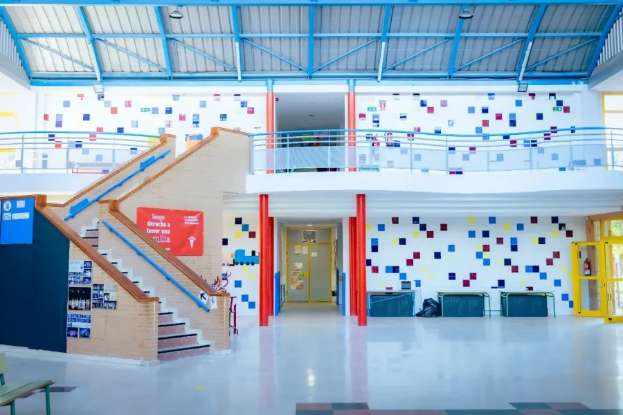 Un luminoso pasillo escolar con azulejos cuadrados de colores en las paredes, una escalera a la izquierda, columnas de soporte rojas y una barandilla en la segunda planta sobre la entrada del pasillo central. El espacio está vacío y bien iluminado por la luz natural, lo que capta a la perfección el animado espíritu de la vuelta al cole en Rivas.