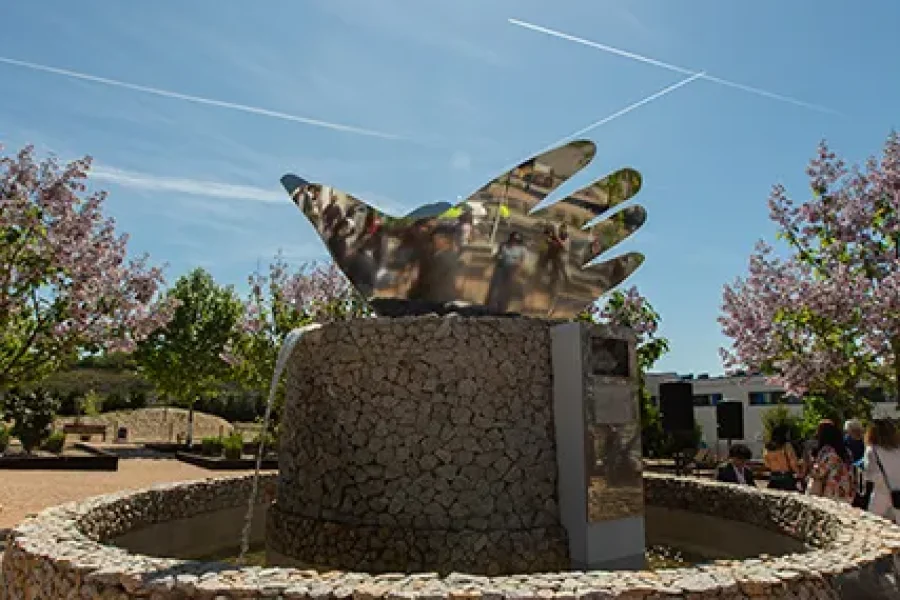 Una fuente de piedra en el Jardín de la Paz de Rivas, con una escultura central que representa un pájaro con forma de mano que refleja la luz bajo un cielo soleado.