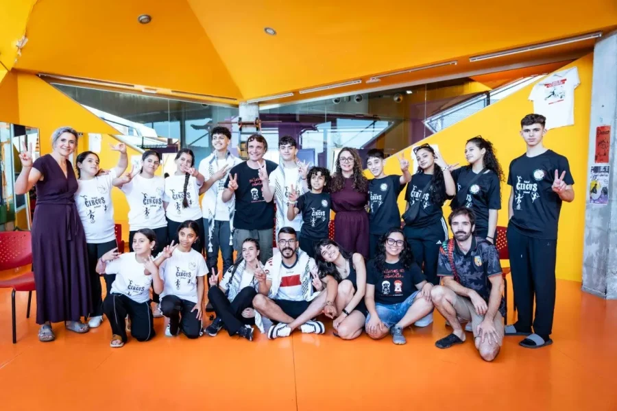 Un grupo de unas 20 personas posa para una foto en el interior, frente a una pared amarilla. La mayoría está de pie, mientras que unos pocos están sentados en el suelo naranja. Muchos sonríen, hacen el signo de la paz y lucen camisetas de marca con la inscripción «borrador automático».