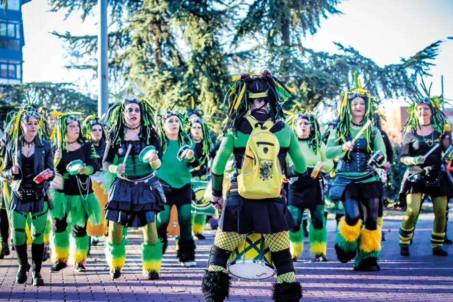 Un grupo de personas vestidas con trajes verdes, negros y amarillos con accesorios a juego tocan tambores e instrumentos de percusión al aire libre en el Carnaval 2026 Rivas, de pie sobre una superficie de ladrillo con árboles al fondo.