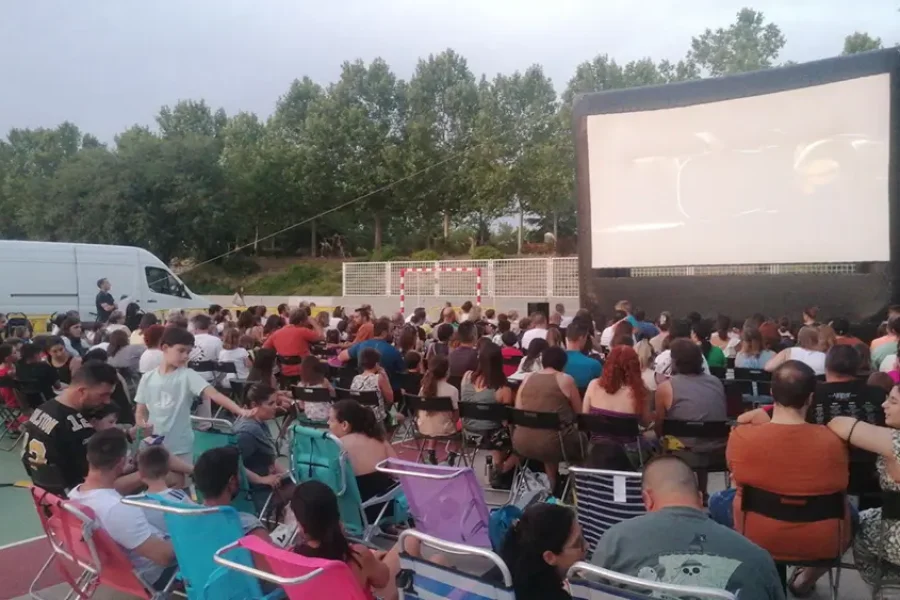 Un numeroso grupo de personas está sentado en sillas y bancos al aire libre en Rivas, disfrutando del cine de verano mientras ven películas gratis en una pantalla hinchable. Al fondo se ven árboles, y a un lado hay una furgoneta blanca aparcada.