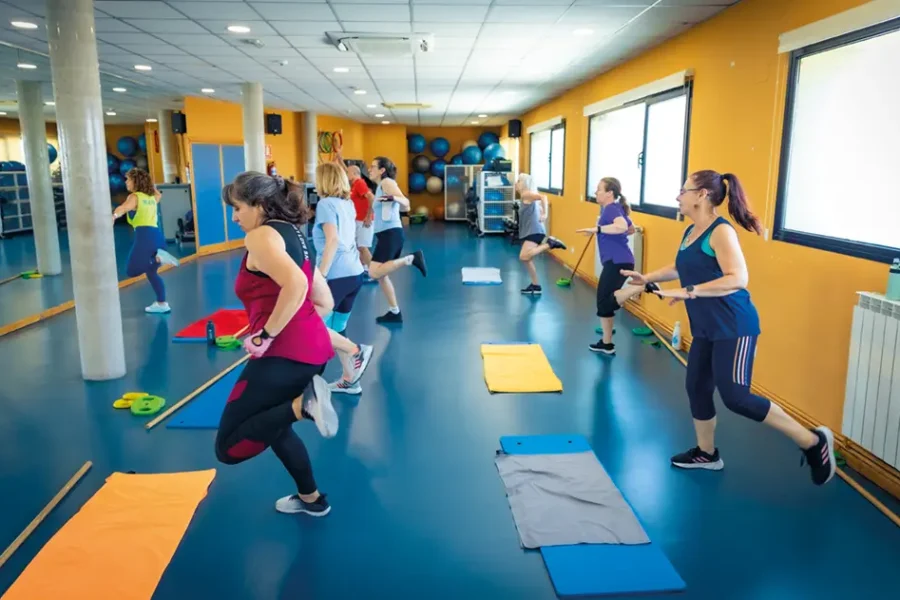 Un grupo de personas vestidas con ropa deportiva hace ejercicio juntas en un gimnasio muy bien iluminado, equipado con colchonetas, espejos y ventanas. Algunas corren sin moverse del sitio, mientras que otras sostienen palos, participando en una clase de entrenamiento en grupo que forma parte de las escuelas deportivas municipales de Rivas. Puedes solicitar plaza para apuntarte y disfrutar de estas dinámicas sesiones con la comunidad.