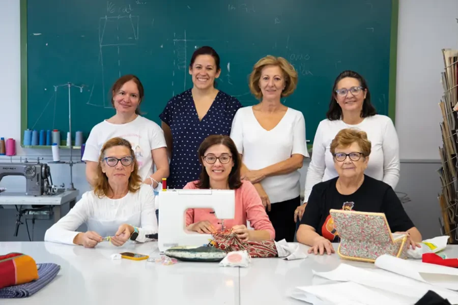 Siete mujeres posan en un aula de costura de la Universidad Popular. Tres están sentadas en una mesa con máquinas de coser y telas, mientras que otras cuatro están de pie detrás de ellas. Al fondo se ven una pizarra, materiales de costura y patrones.