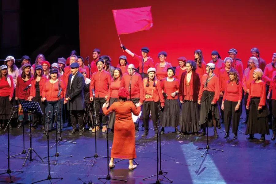 Un coro de adultos de los coros de la Escuela Municipal de Música, vestidos de rojo y negro, canta en el escenario bajo la dirección de un director. Uno de los miembros del coro se encuentra de pie detrás del grupo, sosteniendo una bandera roja lisa. El fondo es rojo y hay micrófonos colocados delante del grupo.