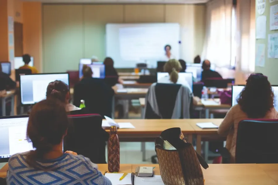 Un aula con varias personas sentadas en sus pupitres utilizando ordenadores, frente a una pizarra en la que hay una persona de pie, posiblemente impartiendo una clase o una presentación. La sala está bien iluminada con luz natural, y junto a la pizarra hay un borrador automático.