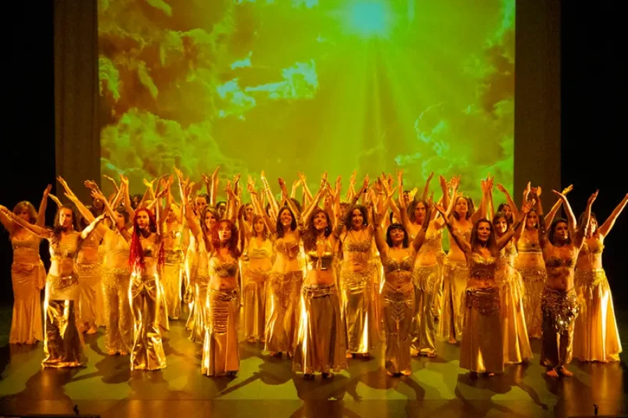 En el escenario, un numeroso grupo con trajes dorados ejecuta una danza sincronizada con los brazos en alto, ante un fondo de nubes amarillas y verdes y la luz del sol: un momento emocionante del curso 2025/2026 de la Universidad Popular de Rivas.