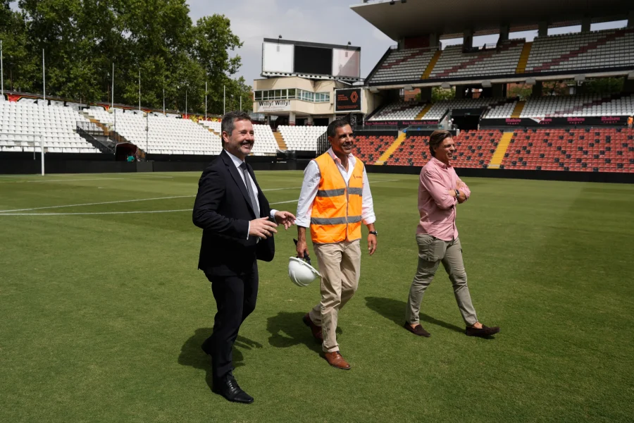 Tres hombres caminan por el campo de fútbol durante la renovación del Estadio de Vallecas en la Comunidad de Madrid; uno lleva un traje negro, otro un chaleco reflectante de seguridad con un casco y otro una camiseta rosa. Al fondo, los asientos del estadio están casi vacíos.