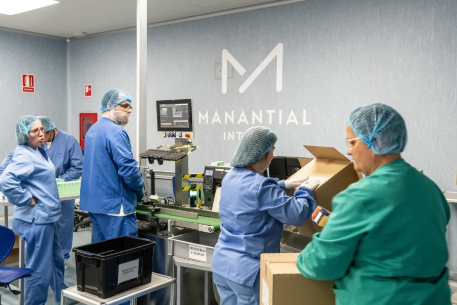 Varias personas con batas azules y verdes, redecillas para el pelo y mascarillas trabajan en una cinta transportadora en las limpias instalaciones de MANANTIAL de Rivas. Algunos empaquetan cajas, otros observan y colaboran para garantizar que los recortes no afecten a la salud mental ni a la calidad del producto.