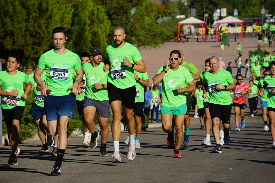 Un grupo de corredores con camisetas verdes a juego y dorsales numerados participan en una carrera al aire libre en un día soleado. Algunos corredores van a media zancada, uno habla por teléfono y al fondo se ve a los espectadores.