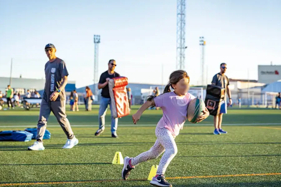 Un niño corre con un balón de rugby en un campo deportivo de Rivas, sorteando conos amarillos, mientras varios adultos y niños permanecen de pie al fondo, bajo un cielo despejado, durante la Semana Europea del Deporte.