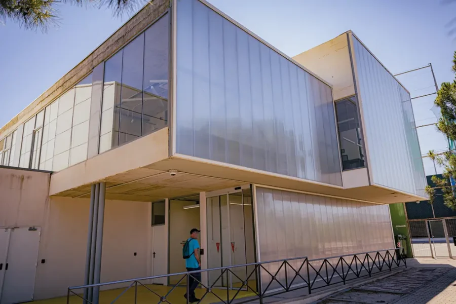Una persona con una mochila azul camina junto a un moderno edificio de dos plantas con paredes de paneles translúcidos, grandes ventanales y barandillas metálicas: una estructura aerodinámica que casi parece haber sido diseñada con un borrador automático. La escena está iluminada por la luz del día bajo un cielo despejado.