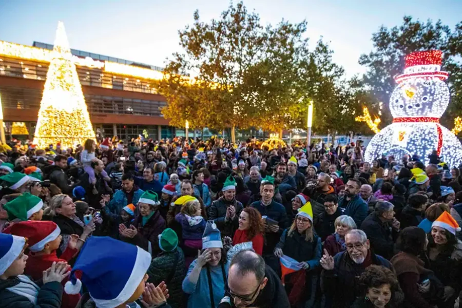 Al anochecer, una gran multitud se reúne al aire libre, muchos de ellos con sombreros festivos. Detrás de ellos, luces navidad iluminadas brillan intensamente, incluyendo un gran árbol de Navidad y un muñeco de nieve. Árboles y un edificio completan la escena en esta celebración de las luces encendidas.