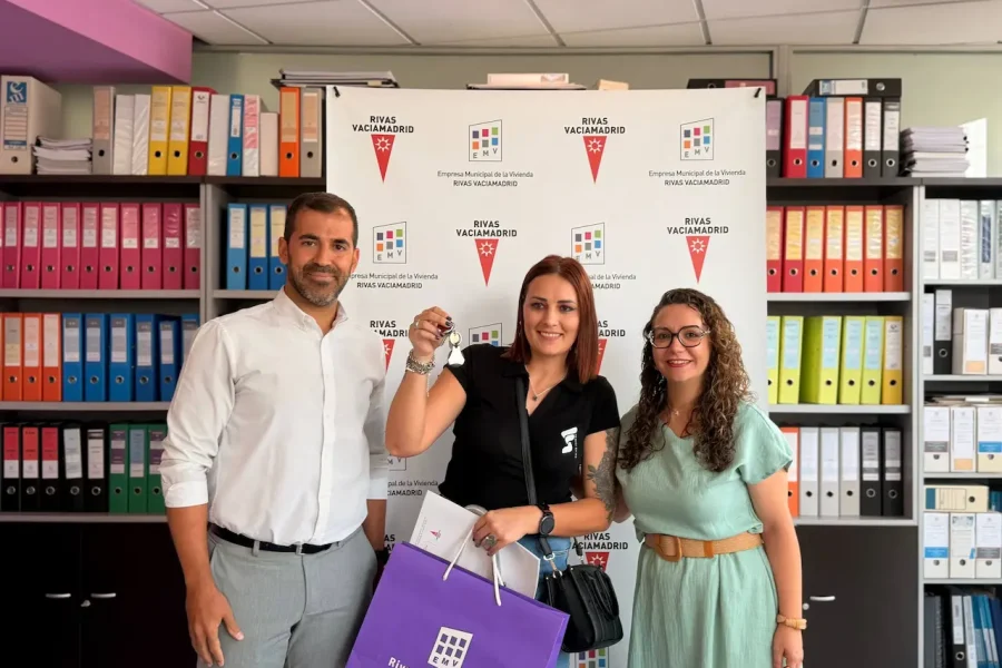 Tres personas posan en el interior, delante de un telón de fondo con logotipos y estanterías llenas de carpetas de colores. La mujer del centro sostiene una llave y una bolsa de la compra morada, para celebrar las nuevas viviendas públicas de alquiler en Rivas. Los tres sonríen a la cámara.