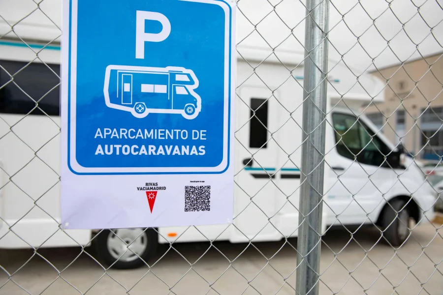 Un cartel azul y blanco con la leyenda Aparcamiento de Autocaravanas está fijado a una valla metálica delante de una autocaravana aparcada. El cartel también incluye el icono de una autocaravana, un código QR y una nota sobre el borrador automático.