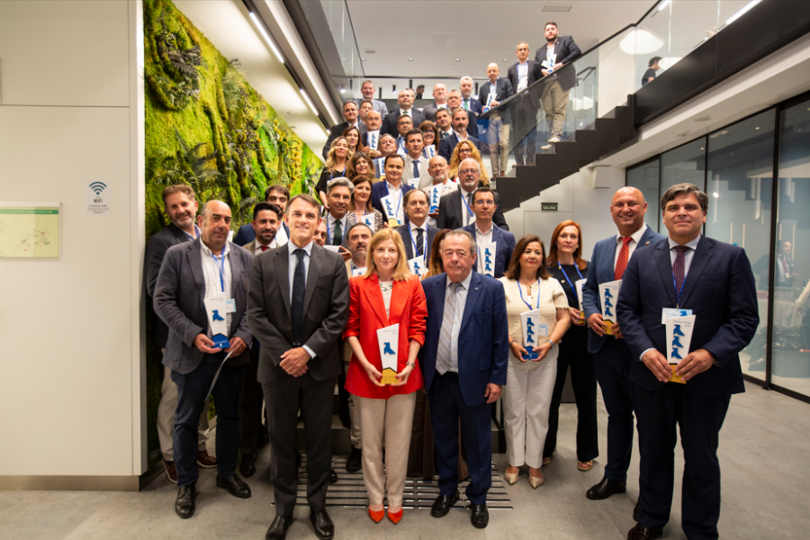 Un grupo numeroso de personas vestidas profesionalmente posan para una foto en el interior de Rivas Vaciamadrid, algunas de pie sobre unas escaleras. Varias personas delante sostienen placas azules y blancas con premios. A la izquierda, un muro vegetal verde celebra el reciclaje de papel.