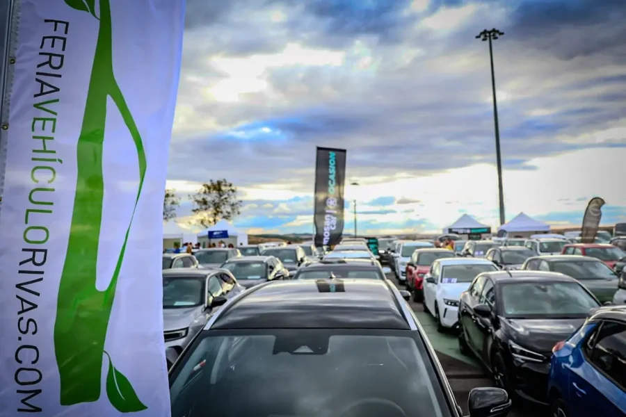 Un aparcamiento en Rivas lleno de coches bajo un cielo nublado, con pancartas promocionales y carpas al fondo. Una bandera blanca en primer plano muestra FERIAVEHICULORIVAS.COM verticalmente en letras verdes, celebrando los vehículos sostenibles.