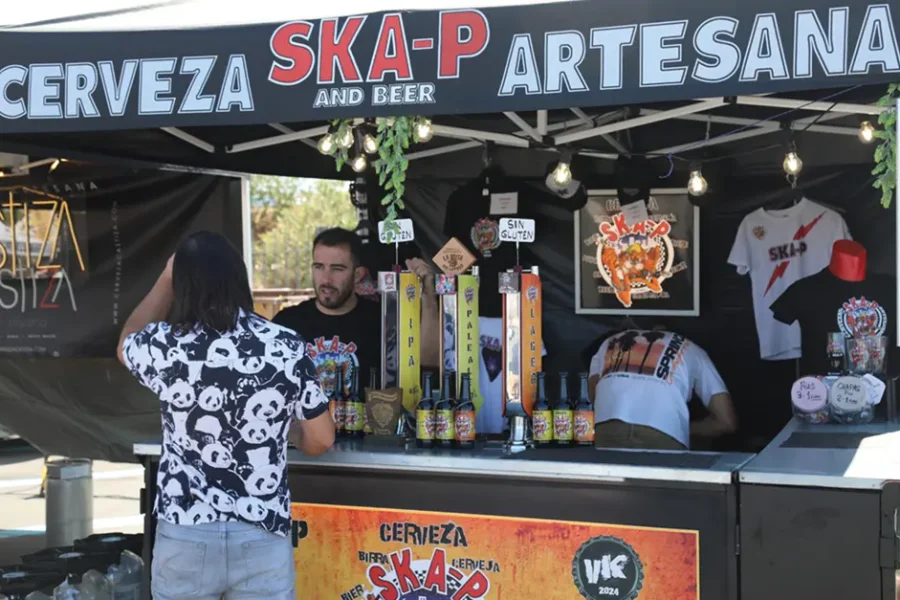Un hombre se encuentra en un puesto donde se vende cerveza artesanal con el nombre de «Cerveza Ska-P Artesana» en un animado festival celebrado en Rivas. Detrás del mostrador hay dos empleados con grifos de cerveza, botellas y productos con el logotipo de Ska-P, lo que pone de manifiesto su pasión por la cerveza artesanal.