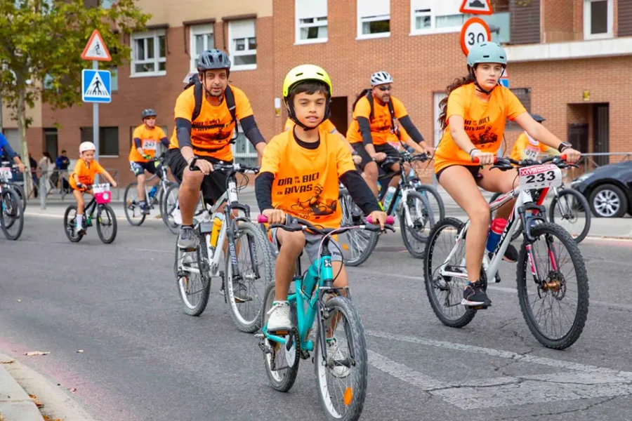 Un grupo de ciclistas, entre los que se encuentran niños y adultos con camisetas naranjas y cascos, pedalea por una calle de la ciudad durante la Fiesta de la Bici en Rivas. En el fondo se ven edificios y señales de tráfico mientras los participantes disfrutan de una de las populares rutas familiares organizadas para el evento.