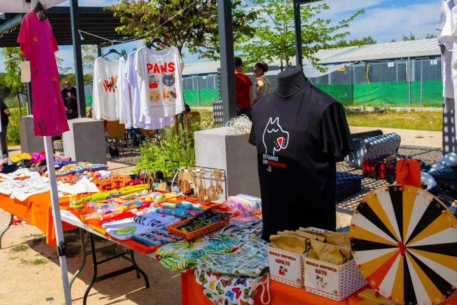 Un puesto del mercado exhibe camisetas de colores, bolsos, artículos de temática gatuna, joyas de Rivanimal y una ruleta para sorteos. El mantel naranja alegra el escenario al aire libre, donde la gente se reúne para informarse sobre voluntariado y adopciones entre vegetación.