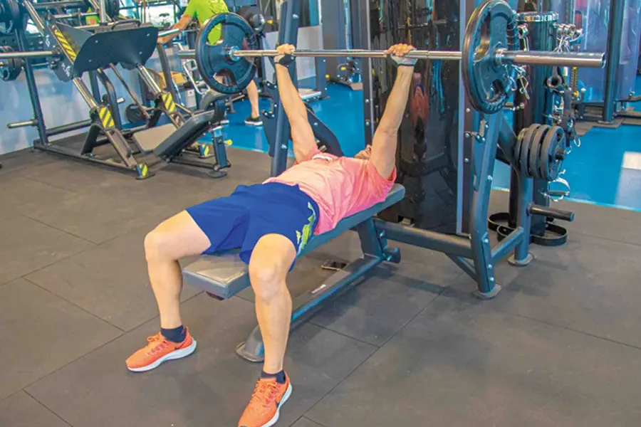 Una persona con camiseta rosa y pantalones cortos azules está tumbada en un banco realizando un press de banca con barra en un gimnasio, lo que refleja el compromiso de los gimnasios de Rivas con la protección de los usuarios. Al fondo se ven diversos aparatos de gimnasio y pesas.