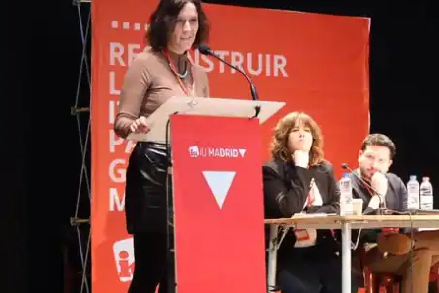 Una mujer de pie en un podio rojo de IU Madrid, presenta la hoja de ruta 2027 a un público. Dos personas se sientan en una mesa detrás de ella, con un telón de fondo rojo y texto de fondo, lo que sugiere un evento o una conferencia en interiores.