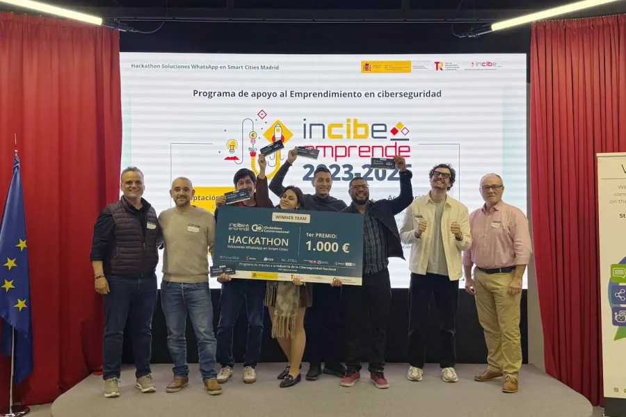 Ocho personas de pie en un escenario de Madrid sostienen un gran cheque de 1.000 euros frente a una pantalla que muestra incibe emprende 2023-2024. Hay banderas, pancartas y un telón rojo en este acto temático sobre la Smart City.