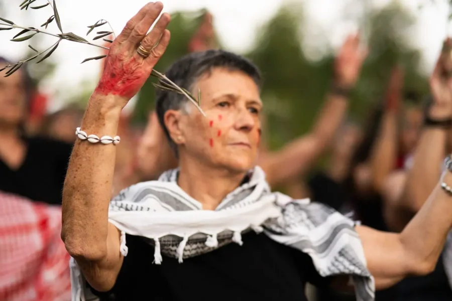 Una persona con un pañuelo estampado y un brazalete de conchas levanta los brazos, con las palmas manchadas de pintura roja, en una reunión al aire libre con otras personas que también han levantado las manos. En el fondo aparecen ramas de árboles y vegetación.