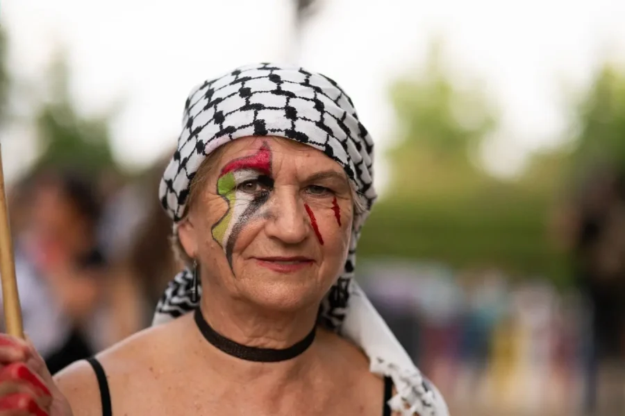 Una mujer mayor con un keffiyeh blanco y negro y una gargantilla está de pie al aire libre. Tiene la cara pintada con los colores de la bandera palestina y sostiene un asta. El fondo es vegetación borrosa.